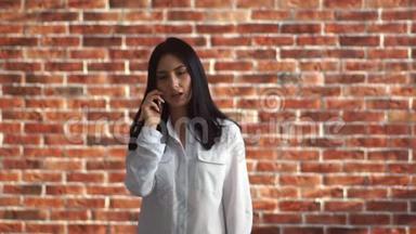 那个女孩在打电话。 一位年轻女子在智能手机上靠砖墙交流